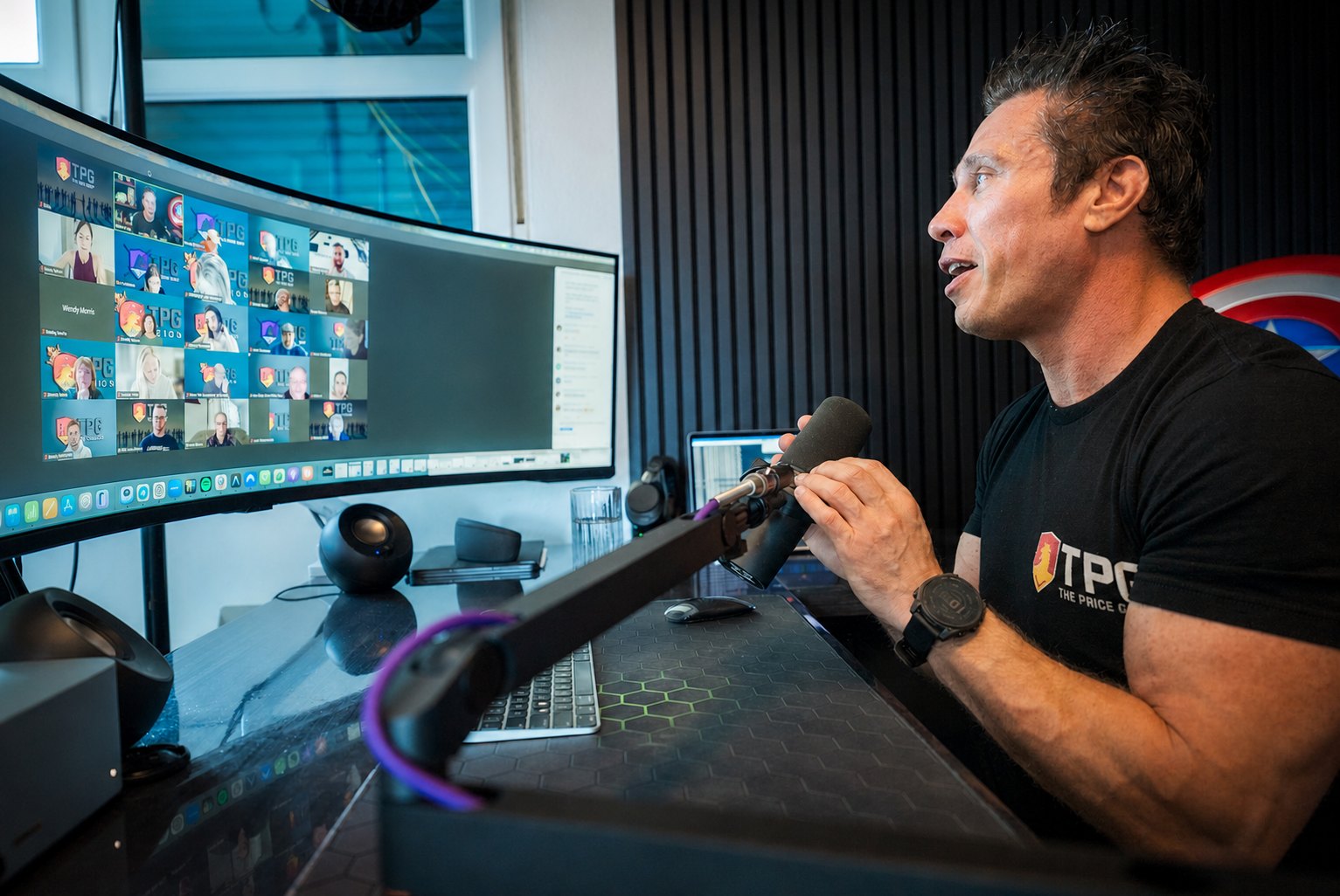 TPG founder David Price leading a virtual TPG team meeting from his workstation, with agents on a large curved monitor and a studio microphone in front of him